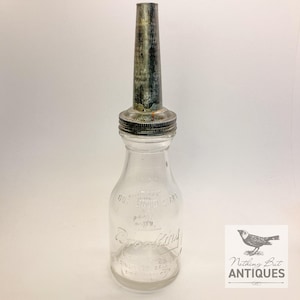 May include: A clear glass oil bottle with a metal spout and a threaded metal cap. The bottle features embossed text that reads Brooking, One Liquid Quart, and Cincinnati, Ohio. The metal spout has a weathered, dark gray finish with visible patina.