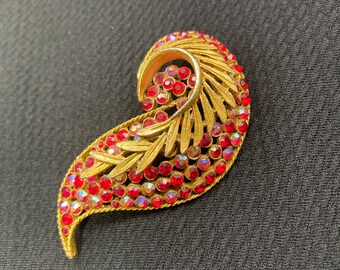 Pode incluir: Um broche dourado em forma de folha estilizada, adornado com strass vermelhos. O broche é uma peça vintage com um design clássico.