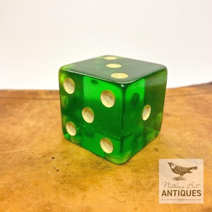 May include: A translucent emerald green six-sided die featuring rounded corners and circular indentations painted in a cream color. The die displays the numbers one, three, and five on the visible faces, with a smooth, polished finish throughout.