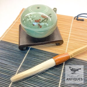 May include: A celadon green ceramic incense burner with a floral design, resting on a dark wooden stand. A calligraphy brush with a wooden handle and white bristles is in the foreground. The image is on a bamboo mat.
