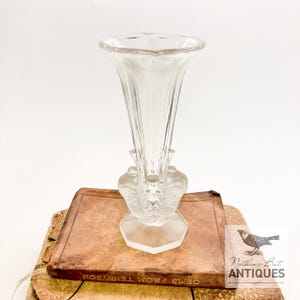 Könnte beinhalten: Eine klare Glasvase mit ausgestelltem Oberteil und dekorativem Sockel. Die Vase steht auf einem alten Buch mit den Worten "GEMS FROM TENNYSON". Die Vase hat ein detailliertes, mattiertes Design am Sockel.