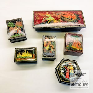 May include: A collection of six antique lacquered boxes with intricate hand-painted scenes. The boxes feature vibrant colors, gold accents, and detailed depictions of figures and landscapes. The collection includes rectangular, square, and hexagonal shapes.