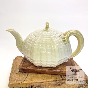 Op de afbeelding: Een lichtgroene keramische theepot met een textuur, geribbeld ontwerp. De theepot heeft een gebogen tuit, een lusvormige handgreep en een deksel met een kleine handgreep. De theepot staat op een houten oppervlak. De tekst "Nothing But Antiques" is zichtbaar.