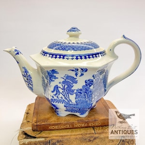 Könnte beinhalten: Eine weiß-blaue Keramik-Teekanne mit Deckel, Ausguss und Henkel. Die Teekanne hat ein blau-weißes Willow-Muster. Die Teekanne steht auf einem Stapel alter Bücher. Die Teekanne ist antik.