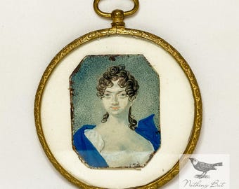 May include: Antique miniature portrait of a woman in a gold-toned frame. The portrait features a woman with dark curly hair and a blue shawl. The frame has a loop at the top for hanging. The portrait is set against a white background.