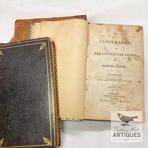Peut inclure: Deux livres anciens reliés en cuir brun avec des accents dorés. Le livre de droite est ouvert pour révéler la page de titre, "The Clockmaker; Or, The Sayings and Doings of Samuel Slick, of Slickville." Le texte continue, "Three from Canada. If here and the Clockmaker speak, we're all alive?" Volume Two, Concord, Boyd and White, Boston, Benjamin B. Mussey, 1850.