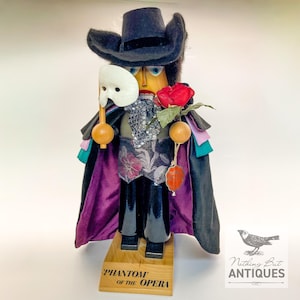May include: A Phantom of the Opera nutcracker figure wearing a black hat and cape with a purple lining. The figure holds a white mask and a red rose, featuring a patterned vest, black trousers, and a wooden base labeled Phantom of the Opera.