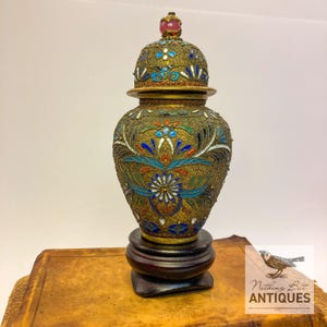 May include: A gold-toned metal jar with a lid, featuring intricate filigree wirework and colorful enamel inlays of blue, green, white, and red. The lid is topped with a small pink stone finial, and the jar rests on a dark wooden circular base.