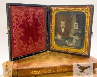 May include: An antique framed photograph of a man and woman, displayed in an open, hinged case. The case has a red velvet interior with an ornate design. The photograph is set on a stack of old books.