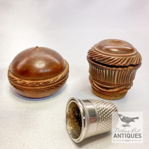 Puede incluir: Tres artículos de costura antiguos: un dedal de plata con superficie texturizada y dos pequeñas cajas de madera tallada intrincadamente. Las cajas son marrones con detalles decorativos tallados. Una caja es redonda y la otra cilíndrica.
