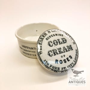 May include: A vintage white ceramic cold cream pot with its lid slightly ajar. The lid displays the text "Cold Cream of Roses" and "120 Fore St. City, London." The pot is marked "Shippam's, Chichester."