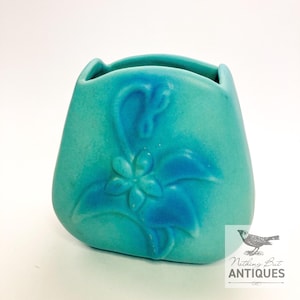Puede incluir: Un jarrón de cerámica turquesa con un diseño floral. El jarrón tiene una forma redondeada con una abertura ancha y un patrón floral en relieve en un tono azul más oscuro. Es una pieza vintage.