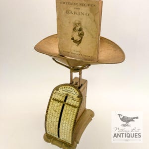May include: Antique brass scale with a vintage cookbook titled "Excellent Recipes for Baking" resting on the scale's tray. The scale features an "Imperial Computing Confectionery Scale" with measurements in pounds and ounces.