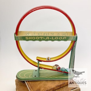 Op de afbeelding: Vintage "Shoot-A-Loop" spel met een rood, geel en groen kleurenschema. Het spel heeft een cirkelvormige lus met een scorebalk en de tekst "SHOOT-A-LOOP". Er zijn verschillende knikkers op de baan te zien. De basis is groen met gebogen lijnen.