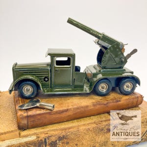 May include: An olive green vintage toy truck with a large cannon mounted on the back. The truck has four wheels and a small key is placed in front of it. The toy is sitting on top of a vintage book.