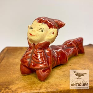May include: A glossy ceramic figurine of a reclining elf with a cream-colored face and pointed ears, wearing a deep red jacket with textured folds. The elf has painted black eyes and a small red mouth, with its body stretched out horizontally.