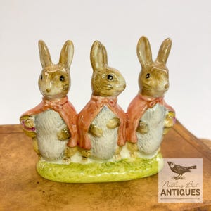 May include: A ceramic figurine featuring three brown and white rabbits standing side by side on a green base. Each rabbit wears a pinkish-red cape. The figurine includes a small logo in the corner with a bird illustration and the text Nothing But Antiques.