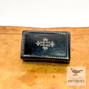 May include: A rectangular black box featuring a silver-toned decorative inlay on the lid. The lid is adorned with a central cross-shaped motif and a delicate scalloped border pattern that runs along the entire perimeter of the top surface.