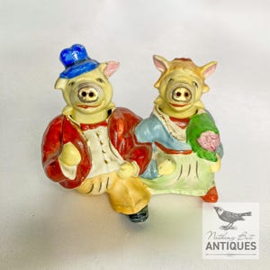 Puede incluir: Un par de figuras de cerdos de cerámica. Un cerdo lleva un sombrero azul y una chaqueta roja, el otro un vestido azul y blanco y sostiene un ramo. Las figuras tienen piel amarilla y están sentadas.
