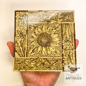 May include: Square, antique box with a detailed sunflower design in the center, surrounded by floral and leaf patterns. The box is in shades of tan and brown, with a decorative border. The box is held in a hand.