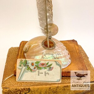 May include: An iridescent abalone shell inkwell with a metal rim, holding a long, mottled brown and white feather quill. The shell is inscribed with "Greetings from Emmett, Idaho" and rests on a vintage book with a floral calling card reading "Lizzie Welly."