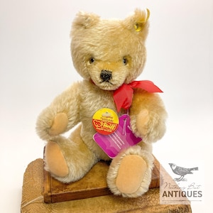 Puede incluir: Un oso de peluche Steiff vintage con pelaje marrón claro, una cinta roja y una etiqueta amarilla. El oso está sentado en una pequeña caja decorativa marrón. La etiqueta dice "Original Teddybär Steiff".