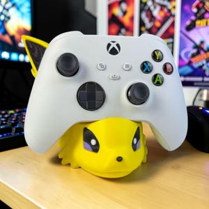 May include: A white Xbox controller rests on a yellow Jolteon-themed stand. The controller has black thumbsticks and colored buttons. The stand is a stylized, yellow figure with black and purple accents, designed to hold the controller.
