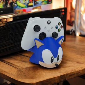 May include: A white Xbox controller rests on a blue Sonic the Hedgehog figure. The controller has black joysticks and colored buttons. The Sonic figure has tan ears and a black nose. The items are on a wooden surface.