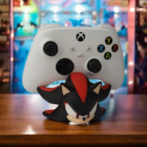 May include: A white Xbox controller rests above a black and red figurine of a video game character. The controller has black buttons and colored letter buttons. The figurine has a white face and red accents.