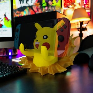 May include: A yellow Pikachu-themed Nintendo Switch dock. The dock features Pikachu's face and arms, with a flame-like design around the base. A Nintendo Switch console is inserted into the dock, with a red and black controller visible.