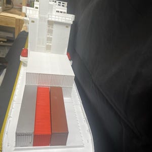 May include: A white model cargo ship with a detailed superstructure and three colorful shipping containers. The containers are silver, red, and brown. The ship has a flat deck and is designed to carry cargo.