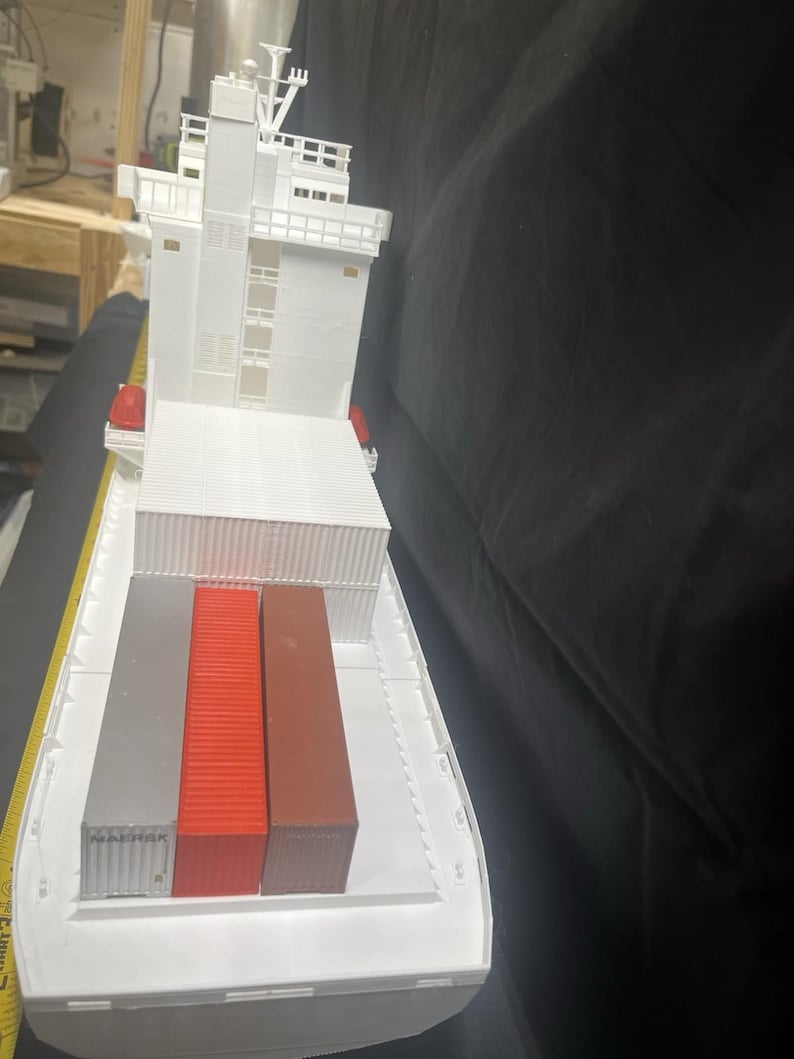 May include: A white model cargo ship with a detailed superstructure and three colorful shipping containers. The containers are silver, red, and brown. The ship has a flat deck and is designed to carry cargo.
