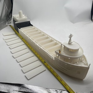 May include: A white 3D printed model of a large cargo ship with a black deck and a small figure standing on the deck. The ship has a long, narrow hull with a rounded bow and a flat stern. The ship is sitting on a white surface with a yellow measuring tape running along the side.