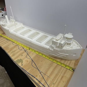 May include: A white model of a cargo ship with detailed features, including multiple cargo holds, a bridge, and masts. A yellow measuring tape is placed next to the ship. The ship model is approximately 30 inches long.
