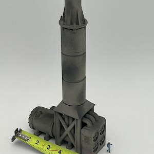 May include: A gray metal model of an industrial smokestack with a base and a series of pipes. The model is approximately 6 inches tall and is being measured with a yellow tape measure.