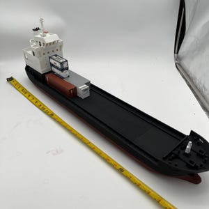 Könnte beinhalten: Ein Spielzeugmodell eines Frachtschiffs mit schwarzem Rumpf, roter Wasserlinie und weißem Aufbau. Das Schiff transportiert Miniatur-Seecontainer. Ein gelbes Maßband liegt neben dem Schiff und zeigt die Maße in Zentimetern an.