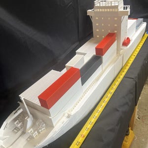 May include: A white model cargo ship with red, black, and white shipping containers. The model is measured with a yellow tape measure. The ship has a detailed bridge and deck features.