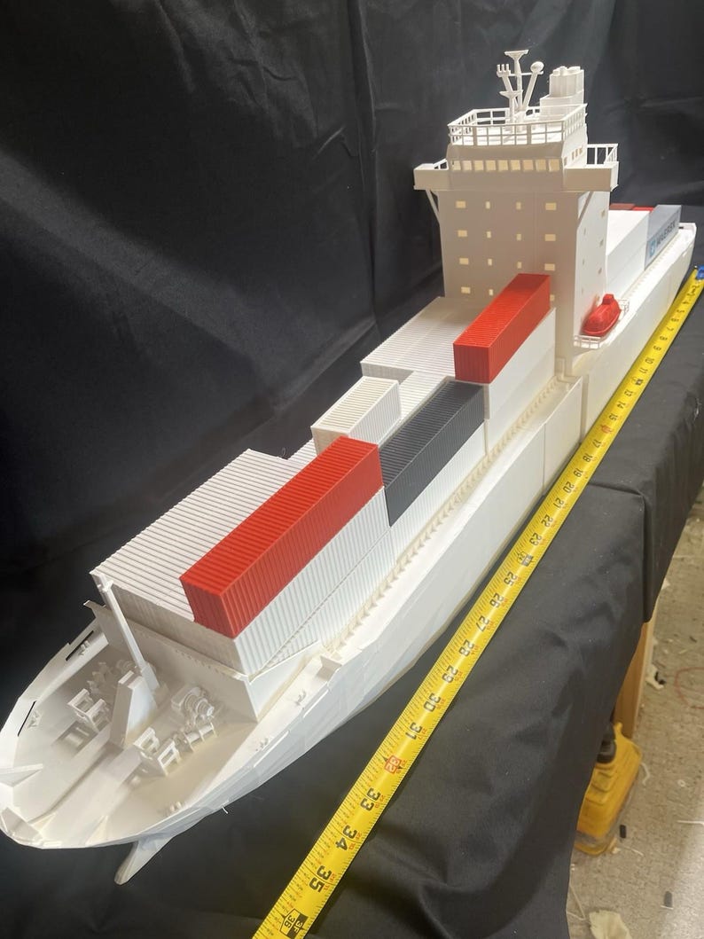 May include: A white model cargo ship with red, black, and white shipping containers. The model is measured with a yellow tape measure. The ship has a detailed bridge and deck features.