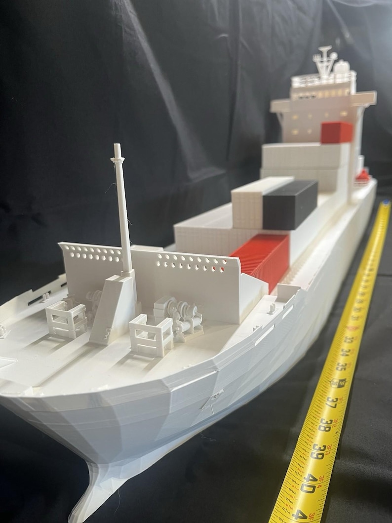 May include: A white model cargo ship with red and black containers. The model is approximately 40 inches long, with detailed features including a mast, deck machinery, and a bridge. The ship is set against a dark background.