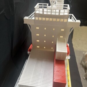 May include: A white and red model cargo ship with detailed structures, including a bridge, deck, and containers. The model has a ruler next to it, showing measurements in inches. The ship's design includes a red container section.