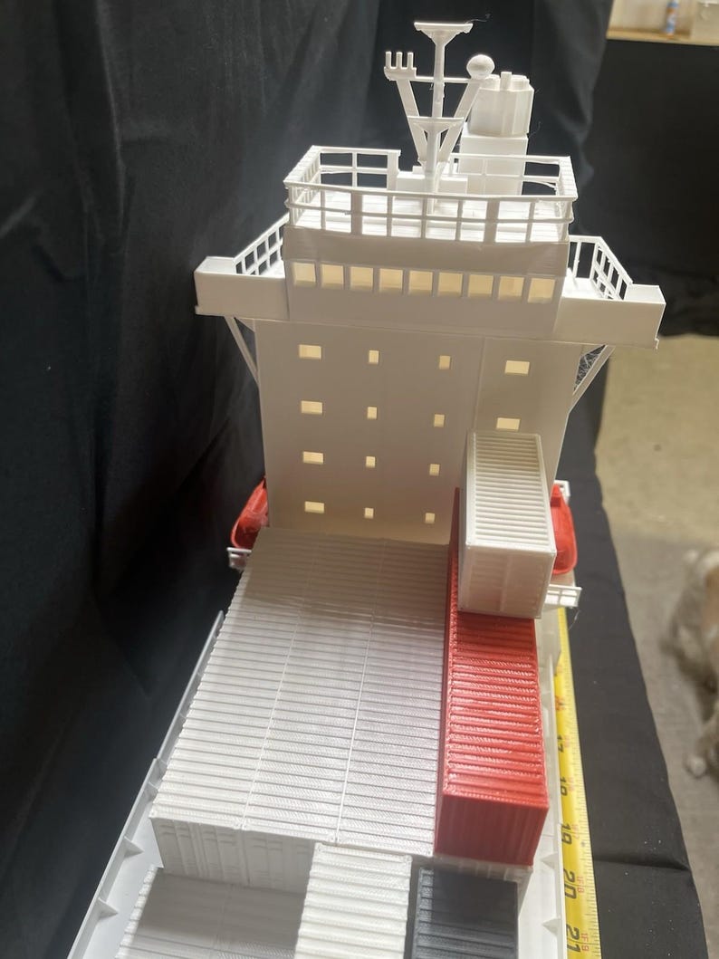 May include: A white and red model cargo ship with detailed structures, including a bridge, deck, and containers. The model has a ruler next to it, showing measurements in inches. The ship's design includes a red container section.