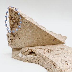 May include: A light pink beaded bracelet with blue flower accents on a rough, beige stone surface.