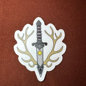 May include: A white sticker with a silver dagger with a yellow sun design on the blade. The dagger is surrounded by two antlers.