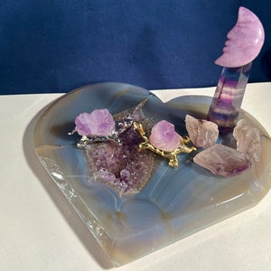 May include: A heart-shaped agate geode with a purple amethyst crystal inside. Two gold turtle figurines are on top of the geode, each holding a purple amethyst crystal. A purple fluorite tower with a purple moon on top is on the right side of the geode.