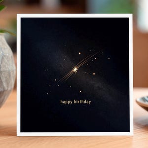 May include: A square birthday card featuring a gold starburst design on a dark blue background. The words "happy birthday" are printed in gold. The card is displayed on a wooden table, with a coffee cup visible.