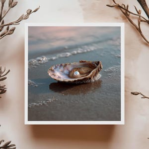 May include: A single pearl sits within an open oyster shell on a sandy beach. The shell is a mix of brown and beige hues, with a gleaming pearl. Soft waves gently touch the shore in the background, under a pale pink sky. The image is framed by slender branches.