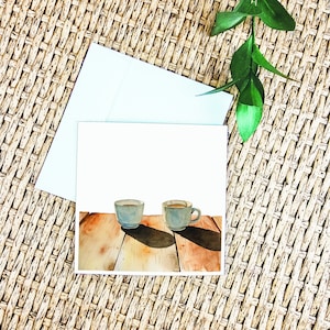 May include: A watercolour painting of two blue coffee mugs on a wooden table. The mugs are filled with coffee and have a light brown colour. The table is made of wood and has a light brown colour.