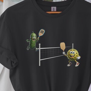 Pickleball T-Shirt: Cartoon Pickle vs Ball Grafik