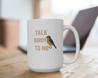 May include: White ceramic mug with the phrase "TALK BIRDY TO ME" in tan letters. A cartoon bird with blue, orange, and red accents is to the right of the text. The mug has a large handle and is on a wooden surface.