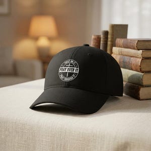 May include: Black baseball cap with a curved peak. The cap has a white circular design with the text "PRAY OVER IT" and "PRAY THROUGH IT". The cap is on a neutral-coloured surface.